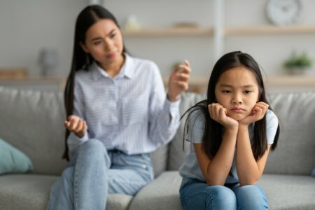 Family misunderstanding concept. Offended asian daughter sitting on couch, turning back to mother