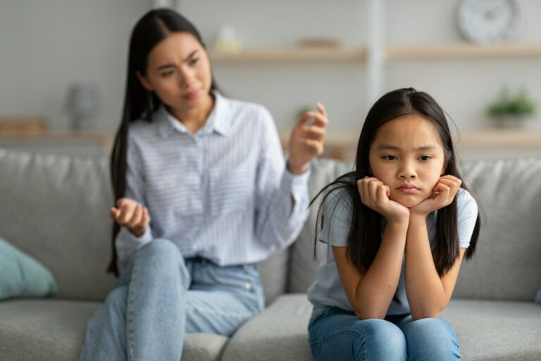 Family misunderstanding concept. Offended asian daughter sitting on couch, turning back to mother
